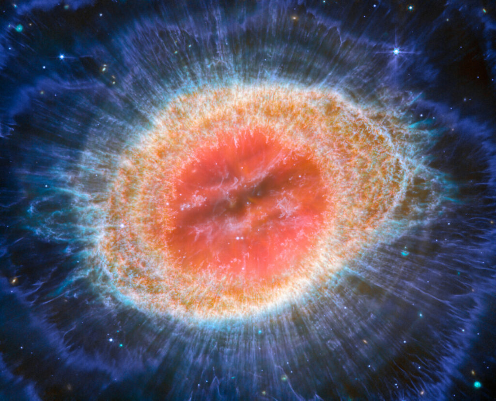 This image of the Ring Nebula appears as a distorted doughnut. The nebula’s inner cavity hosts shades of red and orange, while the detailed ring transitions through shades of yellow in the inner regions and blue/purple in the outer region. The ring’s inner region has distinct filament elements